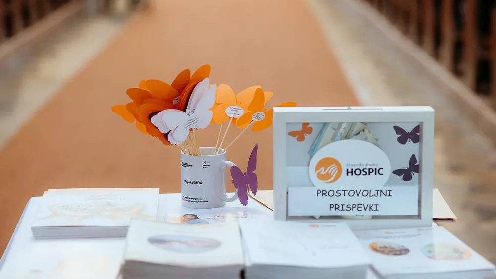 Miza z oranžnimi in belimi papirnatimi metulji, znakom društva in škatlo z napisom Prostovoljni prispevki.