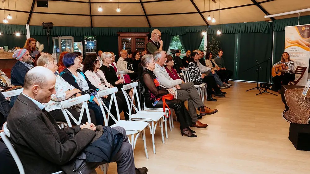 Udeleženci javnega pogovora o smrti in žalovanju sedijo v vrstah v šotoru, ob strani glasbenica s kitaro.
