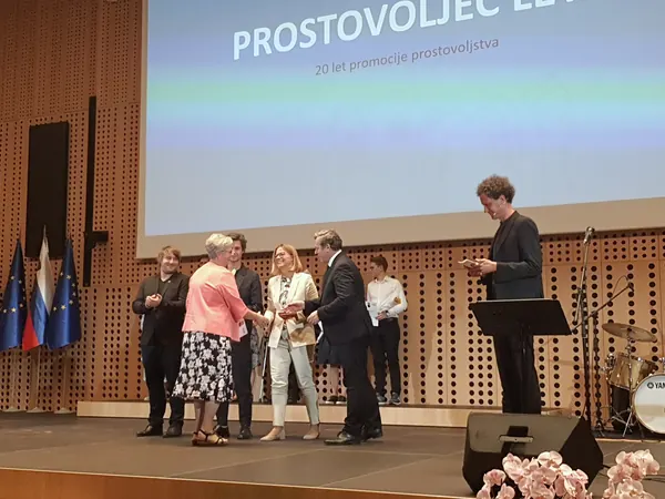 Prejemnica priznanja se na odru rokuje s predstavnikom organizatorja, poleg stojijo drugi udeleženci.