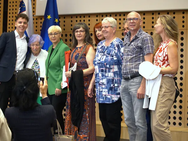 Skupina prejemnikov priznanj stoji skupaj pred oblazinjeno leseno steno, v ozadju državna zastava.