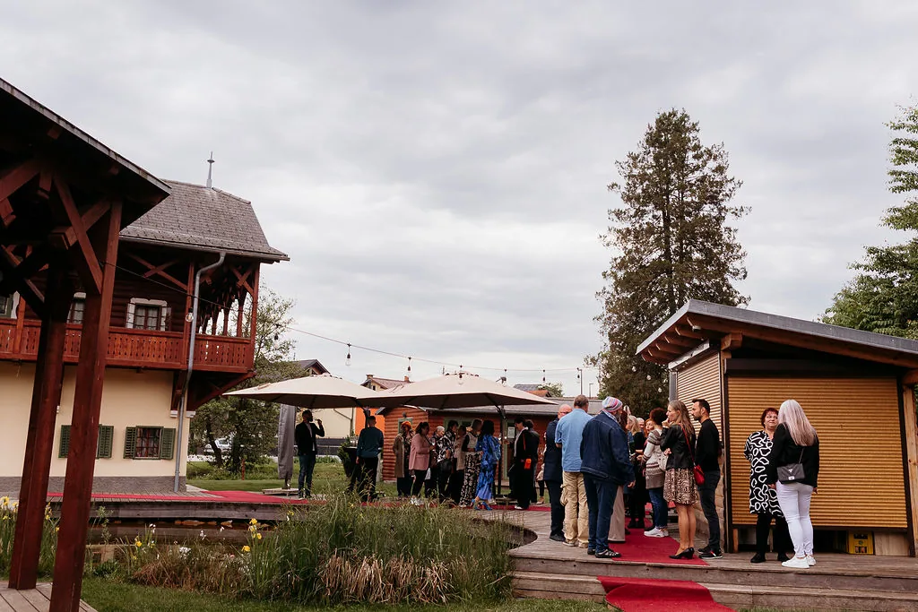 Vrt z lesenimi stavbami in ribnikom, gostje se zbirajo ob vhodu.