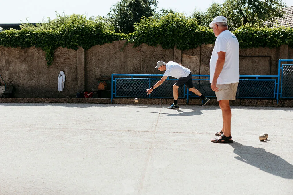 Dva moška igrata balinanje na betonskem igrišču ob zidu z zelenjem.