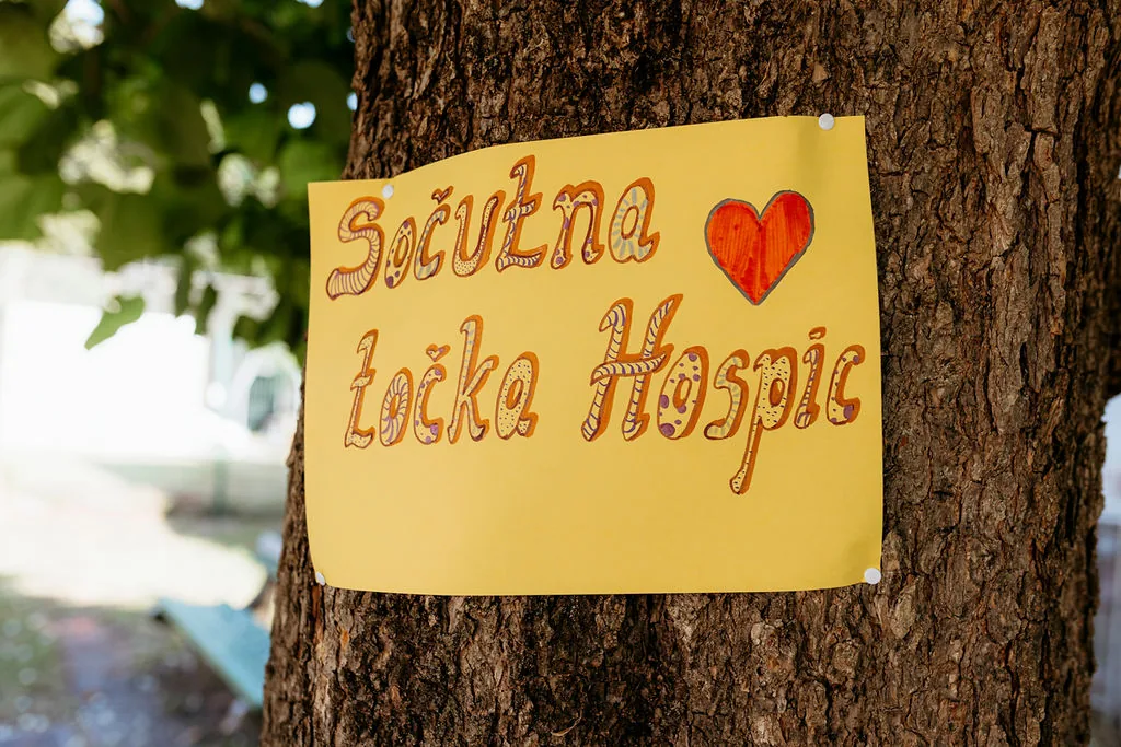 Rumen list z napisom Sočutna točka Hospic in srcem, pritrjen na deblo drevesa.