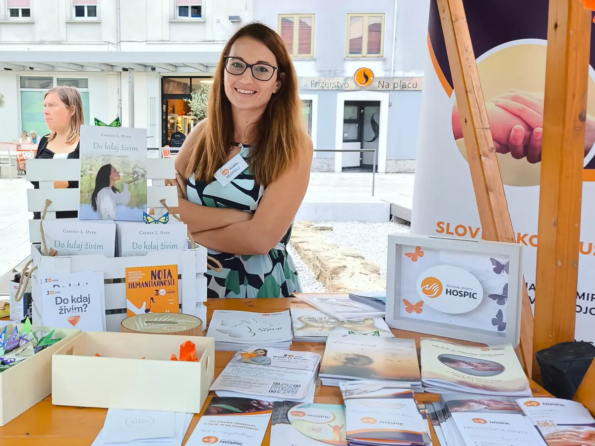 Predstavnica in sodelavka Slovenskega društva Hospic na stojnici ob svetovnem dnevu humanitarnosti, z gradivom društva in papirnatimi metulji na mizi.