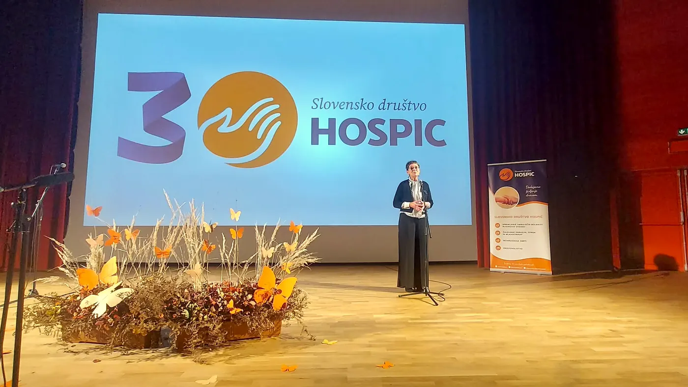 Govornica stoji na odru ob prireditvi ob tridesetletnici društva; za njo je platno z znakom društva, ob odru pa jesenski aranžma s papirnatimi metulji.