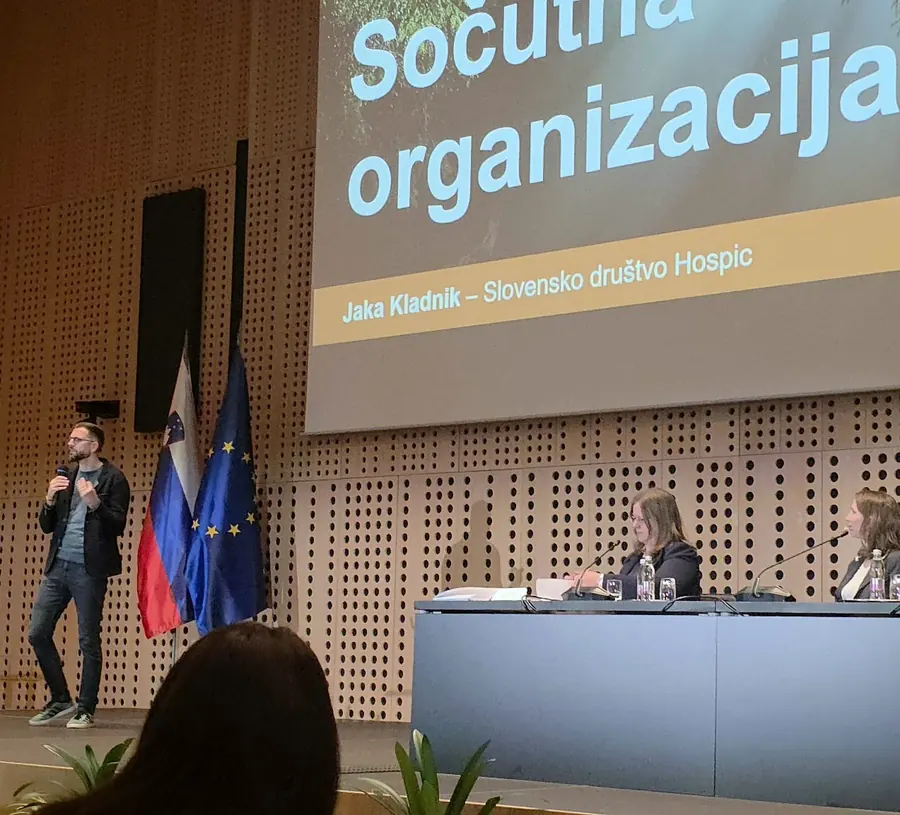 Govornik na odru ob zastavah Slovenije in Evropske unije, nad njim platno s prosojnico Sočutna organizacija, desno za mizo sedita dve osebi.